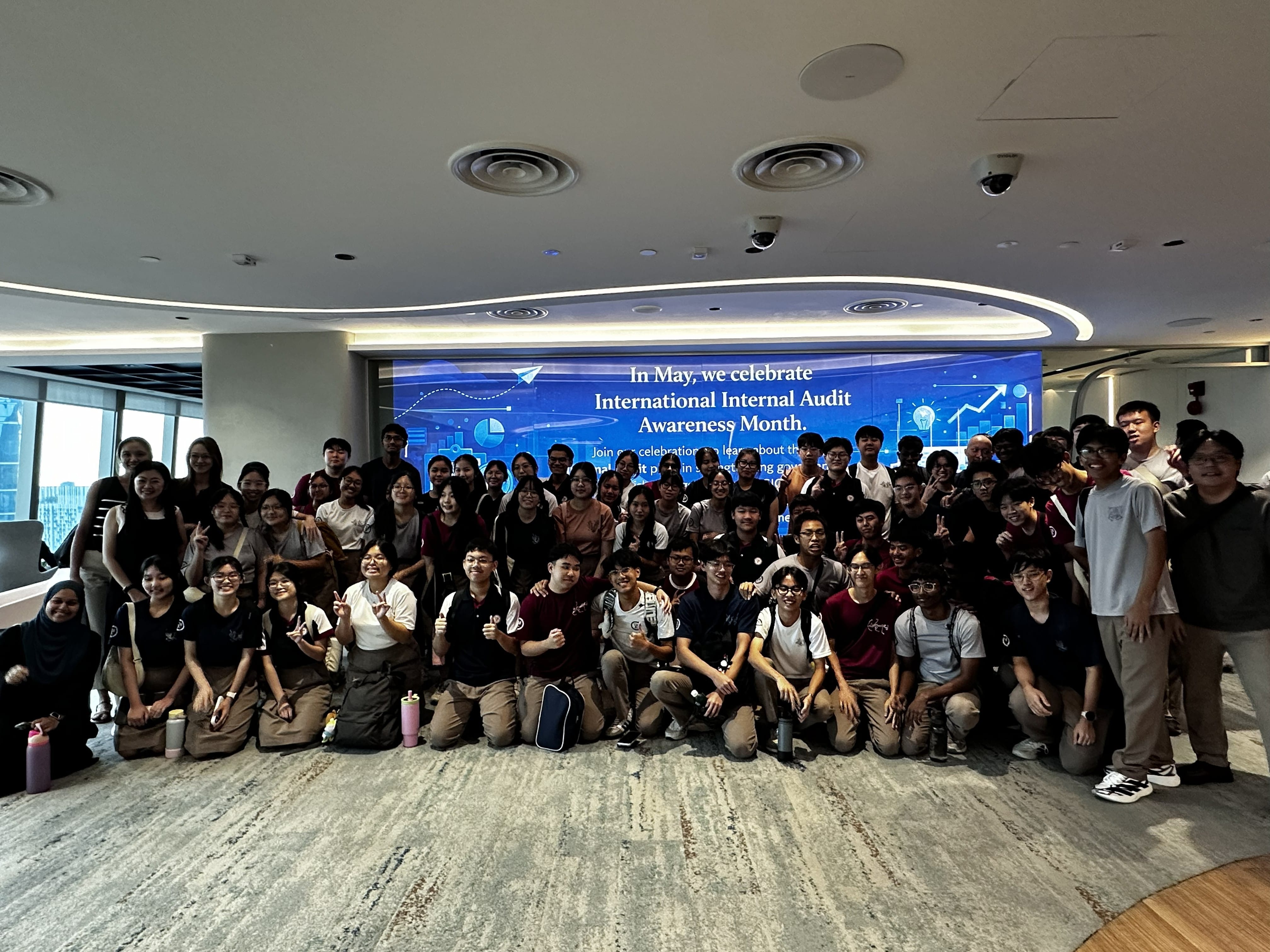 A large group of students and teachers pose together for a group photo inside a modern office space. Behind them is a large blue digital screen displaying the message "In May, we celebrate International Internal Audit Awareness Month."